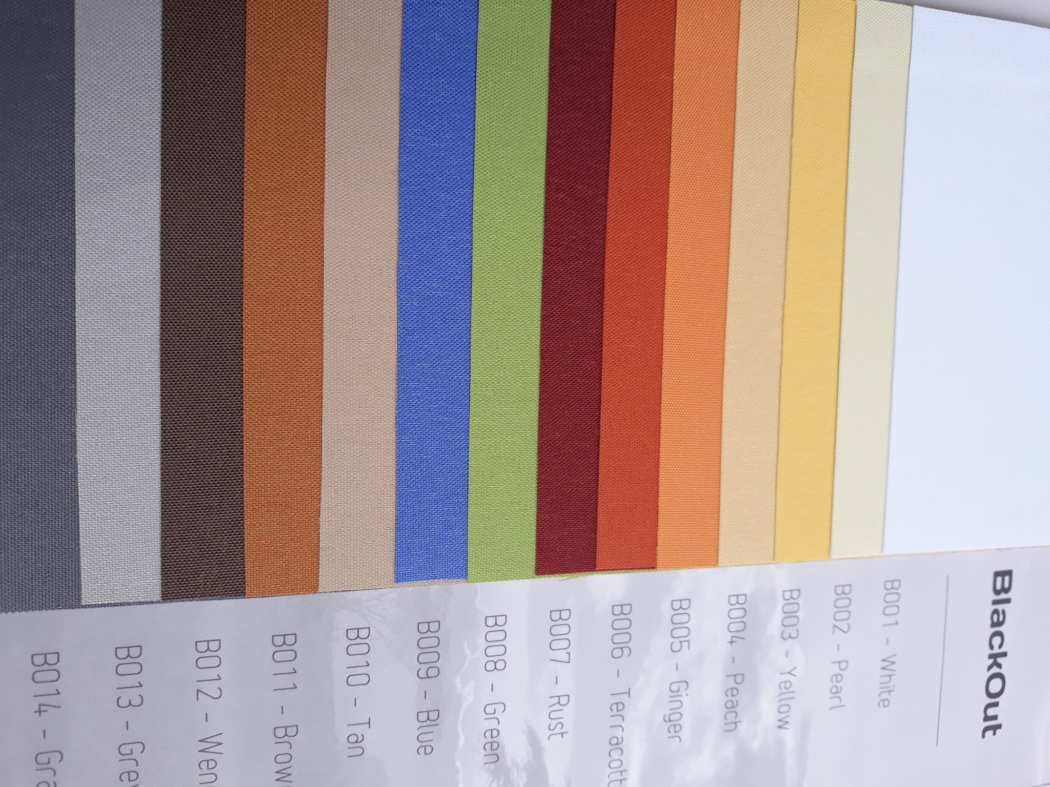 Paleta kolorów tkanin Blackout z oznaczeniami B001-B014, prezentująca próbki materiałów w odcieniach bieli, beżu, żółci, pomarańczy, czerwieni, zieleni, niebieskiego, brązu i szarości.