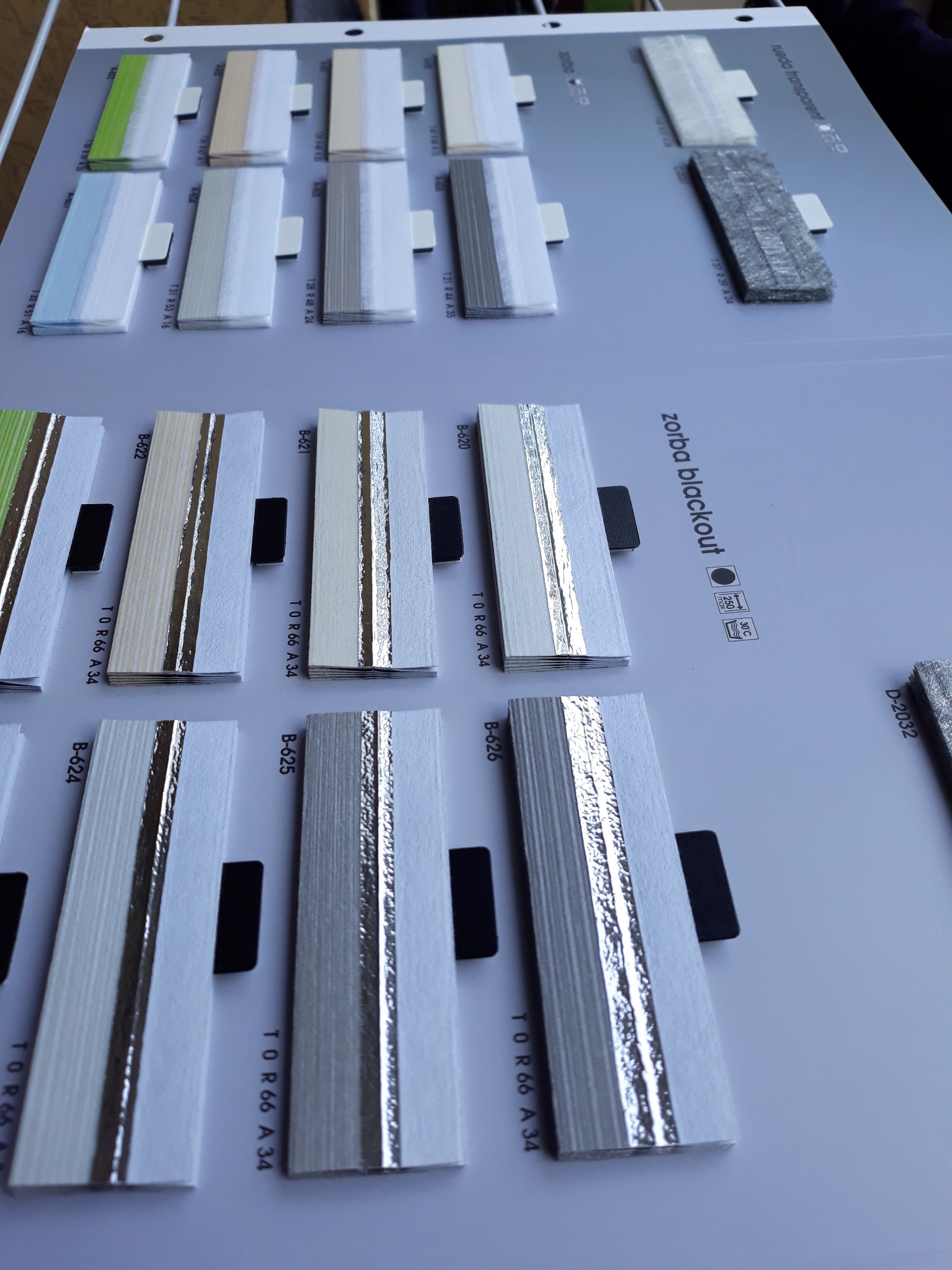 Paleta próbek różnych kolorów i faktur materiałów na rolety lub żaluzje, z widocznymi numerami identyfikacyjnymi i oznaczeniami producenta.