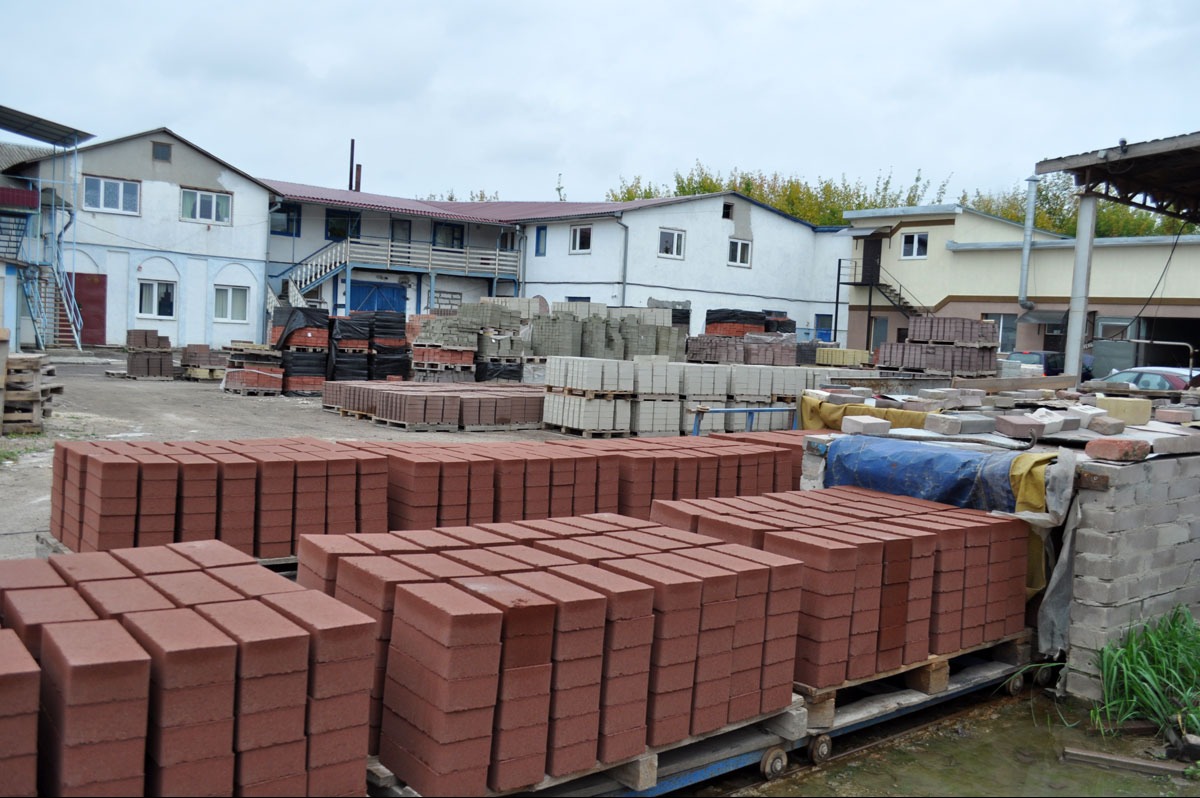 Składowisko kostki brukowej w odcieniach czerwieni i szarości, ułożonej na paletach, z budynkami w tle.