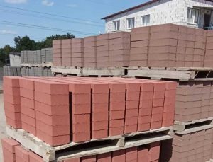 Ułożone na drewnianych paletach, rzędy czerwonej kostki brukowej w składzie budowlanym, budynek w tle.