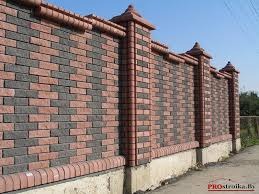 Mur z dwukolorowej cegły klinkierowej, z dekoracyjnymi elementami na szczycie słupków, osadzony na betonowej podmurówce. Widoczny fragment terenu i błękitne niebo.