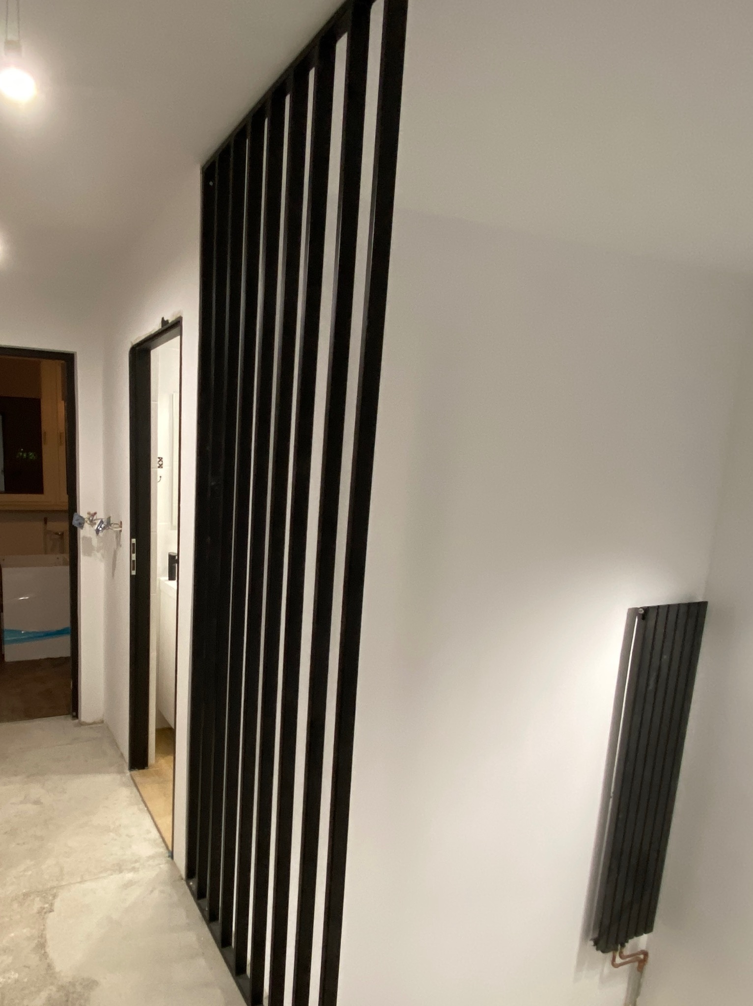 Nowoczesna aranżacja wnętrza: czarne, pionowe lamele oddzielają przestrzeń. Widoczny fragment korytarza i grzejnik na ścianie. Minimalistyczny design.