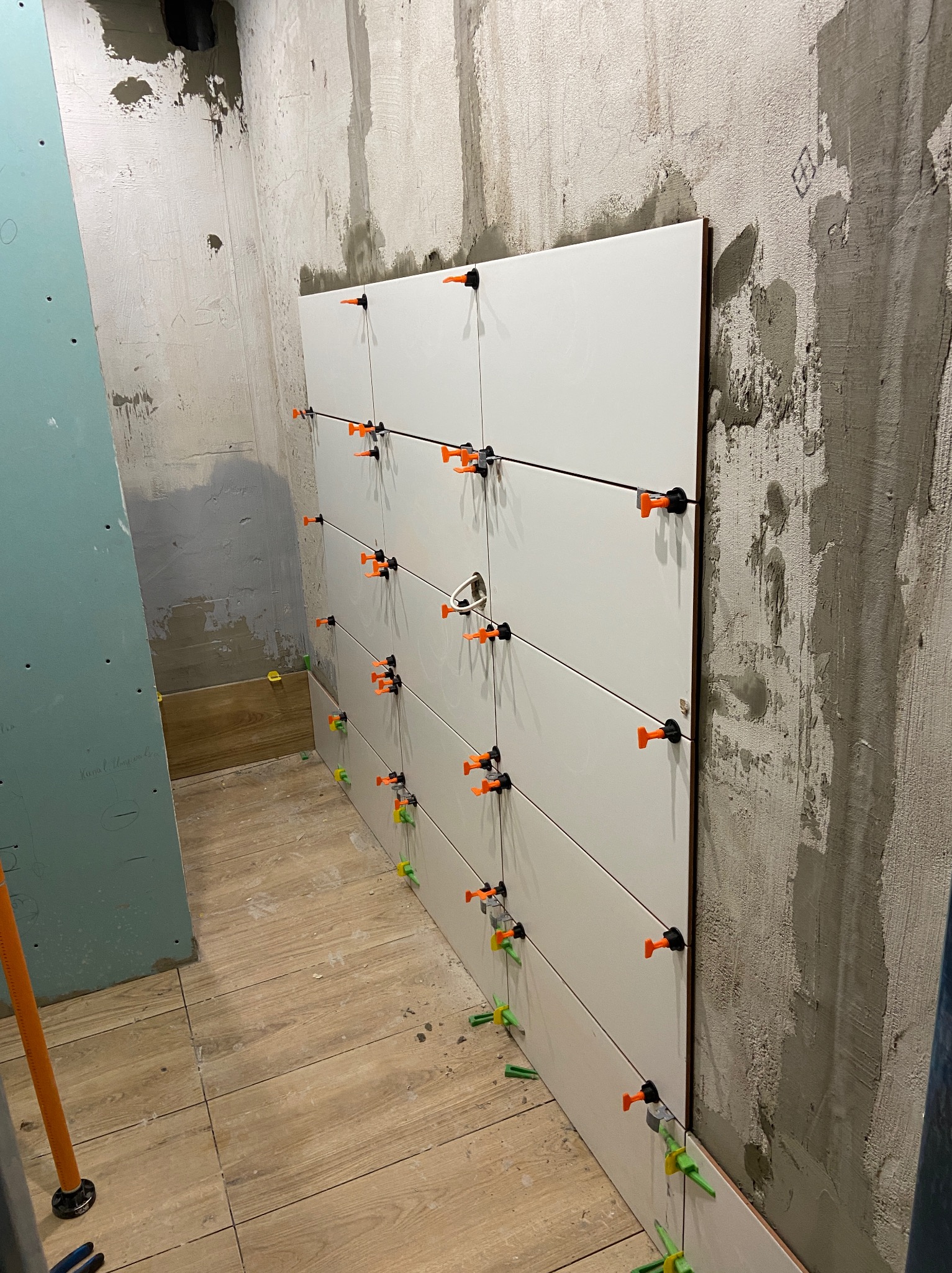 Układanie białych, podłużnych płytek na ścianie z użyciem pomarańczowych i zielonych klipsów poziomujących. Widoczne surowe ściany i fragment podłogi z płytek imitujących drewno.