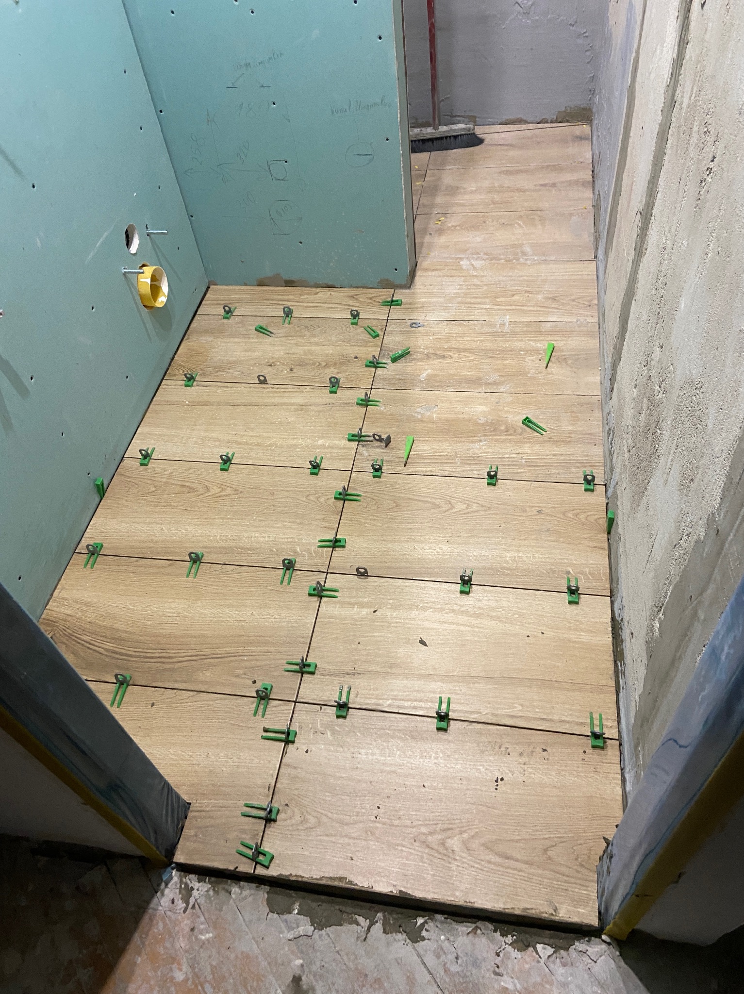 Układanie płytek podłogowych imitujących drewno w łazience, z użyciem zielonych klipsów poziomujących. Widoczne surowe ściany i płyta gipsowa z instalacją.