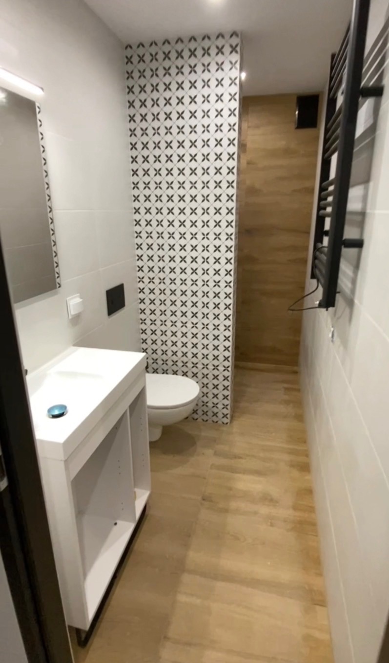 Nowoczesna łazienka po remoncie: biała umywalka z szafką, toaleta z wzorzystą ścianką działową, drewnopodobne płytki podłogowe i czarny grzejnik.
