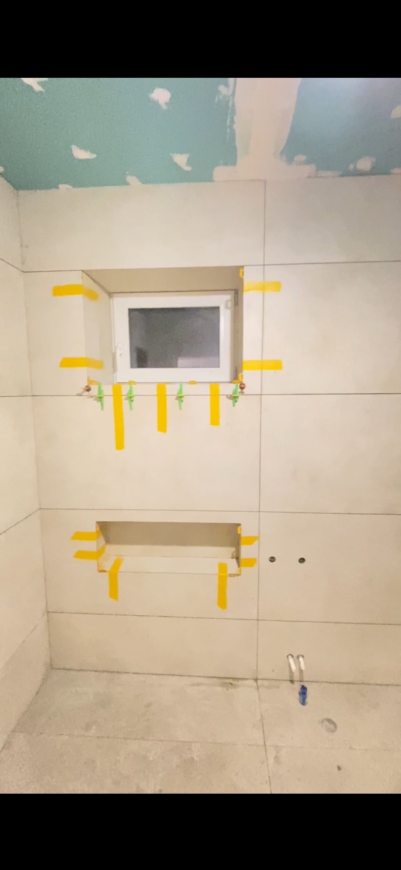 Białe płytki ścienne w łazience, z oknem i wnęką, zabezpieczone żółtą taśmą malarską. Widoczne poziomice i krzyżyki glazurnicze. Sufit częściowo wykończony.