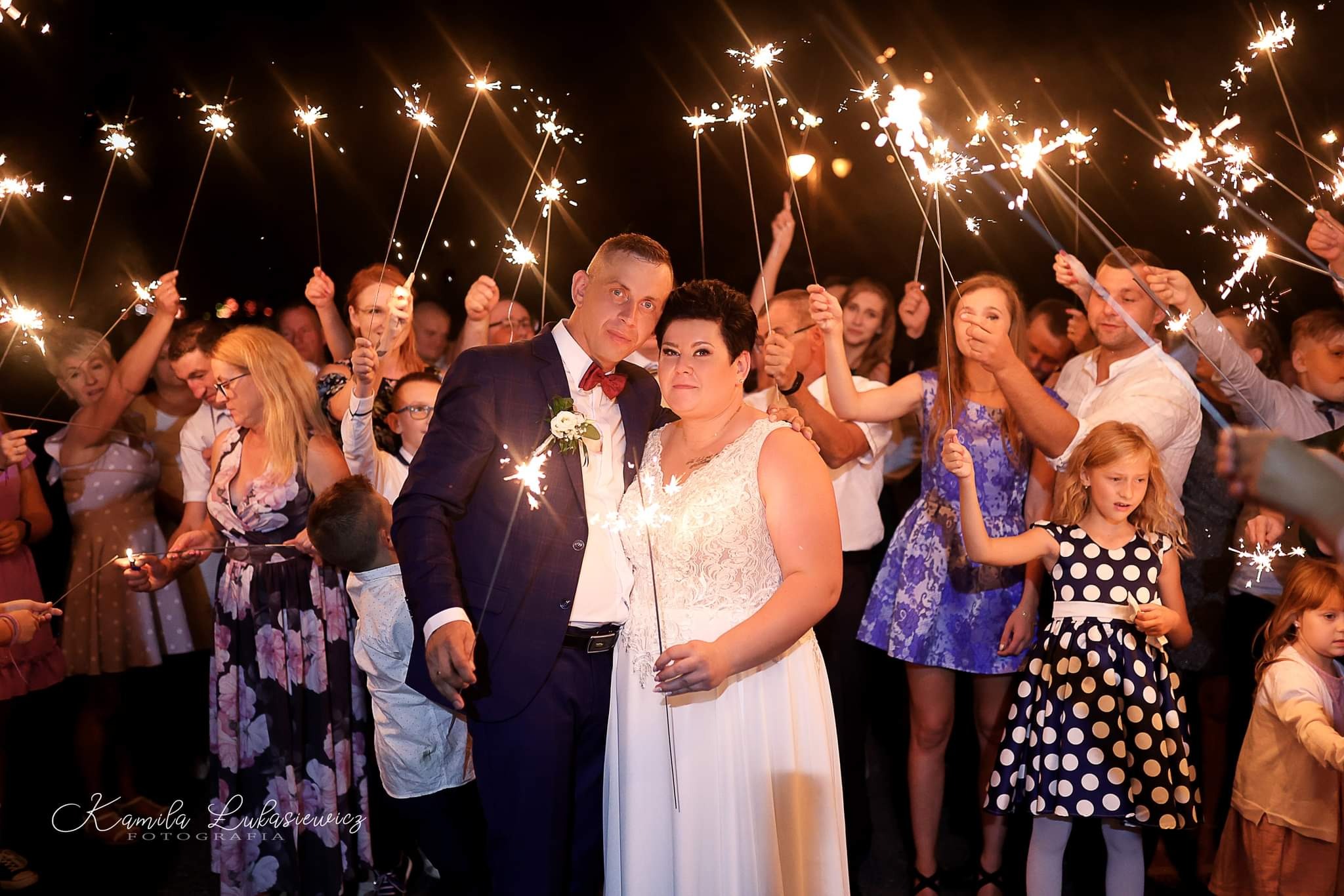 Para młoda otoczona gośćmi weselnymi trzymającymi zimne ognie na tle ciemnego nieba.