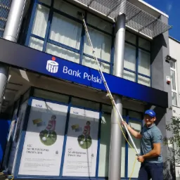 Pracownik w niebieskiej czapce czyści okna budynku banku PKO długą lancą z zamontowaną szczotką, widoczne reklamy Visa na oknach.