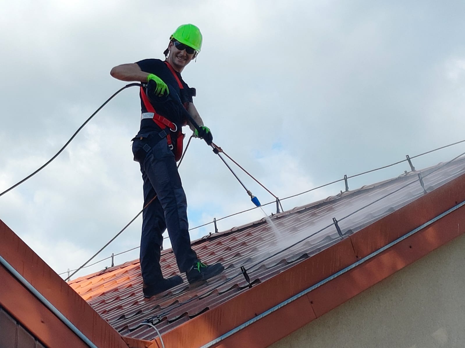 Pracownik w kasku i uprzęży, z uśmiechem, czyści dach z pomarańczowej dachówki za pomocą myjki ciśnieniowej, widoczne chmury na niebie w tle.