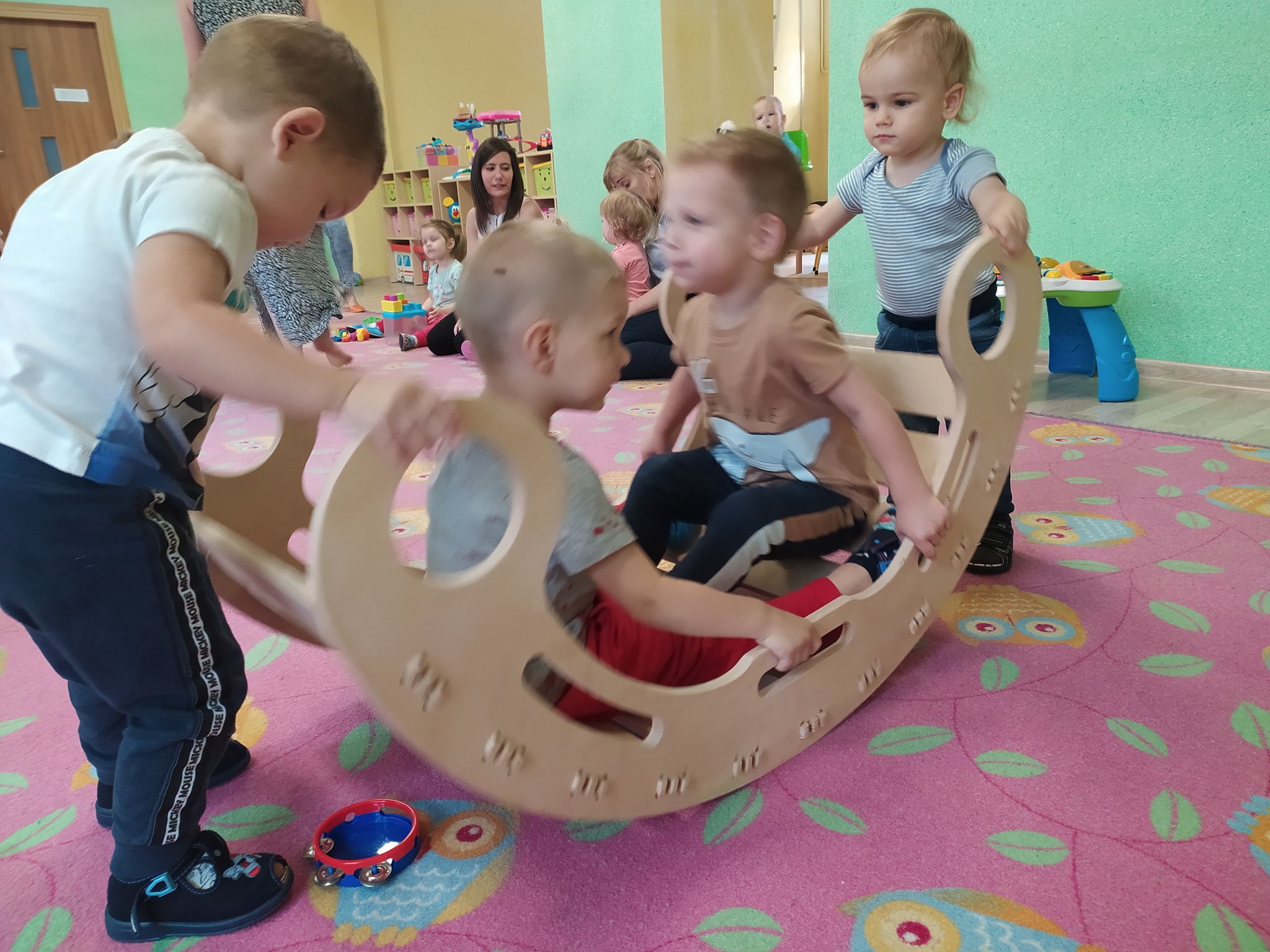 Dzieci bawią się w drewnianej kołysce w sali z zielonymi ścianami i dywanem w sowy, pod nadzorem opiekunek.