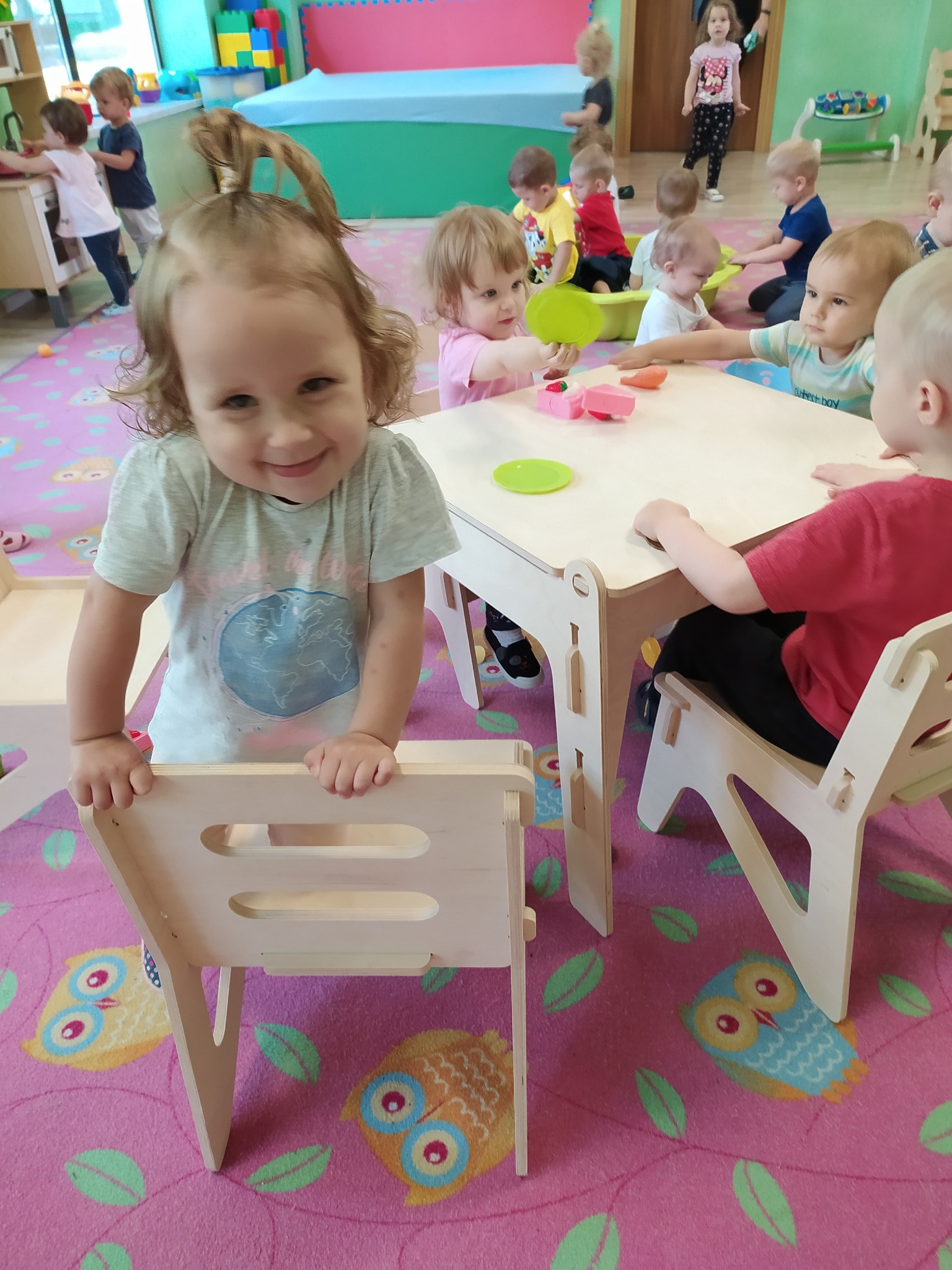 Uśmiechnięta dziewczynka opiera się o jasne, drewniane krzesełko na tle sali zabaw z innymi dziećmi i stolikiem z zabawkami.