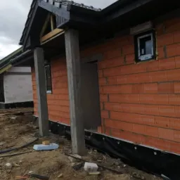 Surowy stan budynku w trakcie budowy: ściany z czerwonej cegły, czarny dach z widoczną więźbą, betonowe kolumny podtrzymujące zadaszenie, okna wstawione, teren wokół budynku nierówny, z widocznymi...