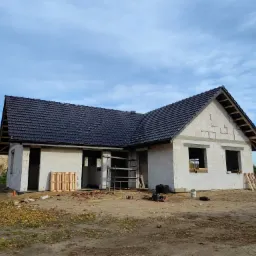 Dom w trakcie budowy z ciemną dachówką, widoczna więźba dachowa, ściany z białych bloczków, rusztowanie i palety w otoczeniu placu budowy.