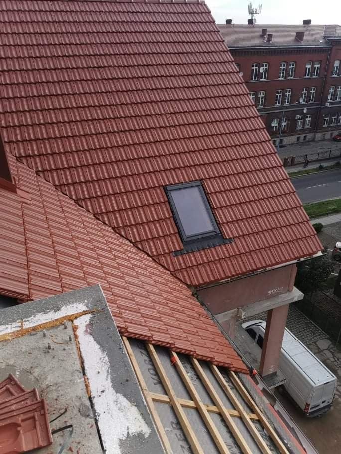 Nowy dach z czerwonej dachówki ceramicznej, widoczne okno dachowe, fragment więźby i budynek w tle.