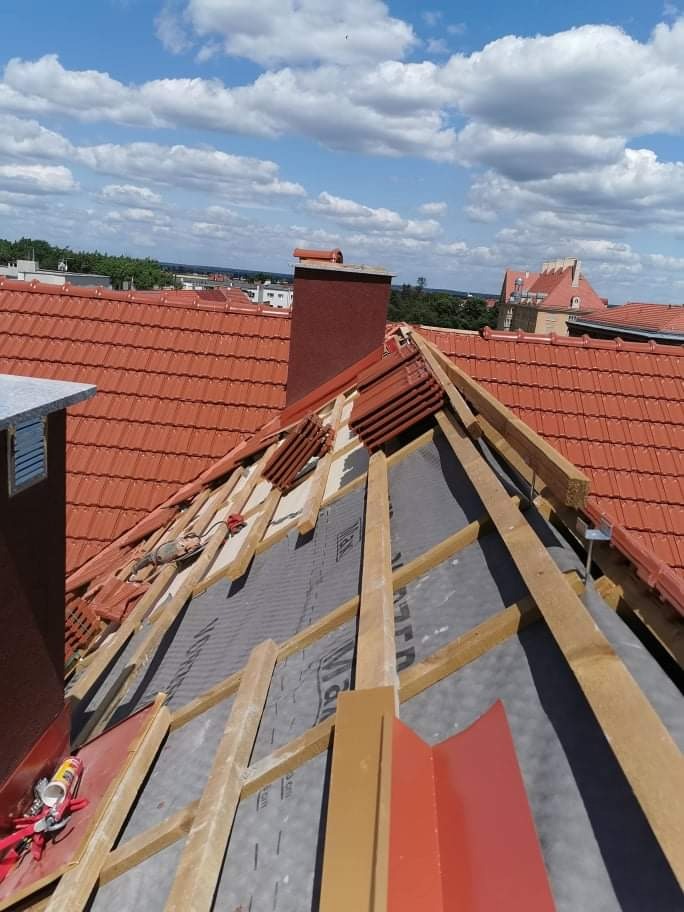 Częściowy widok dachu z czerwoną dachówką w trakcie prac, widoczna membrana, drewniana konstrukcja, komin i narzędzia, na tle błękitnego nieba z chmurami.