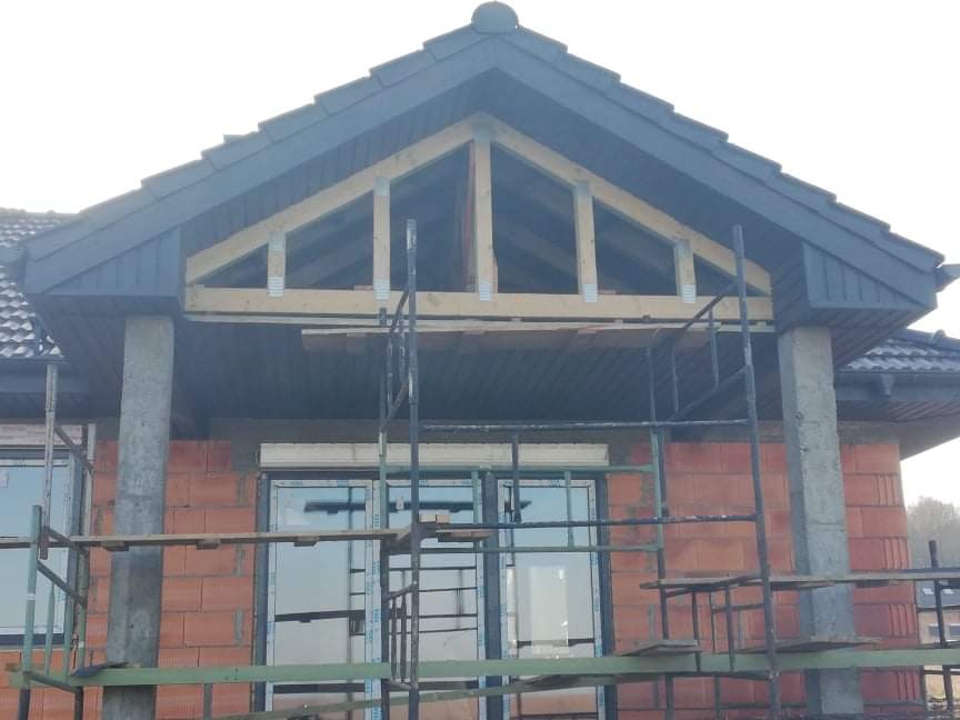 Wznoszenie drewnianej więźby dachowej nad wejściem do domu z cegły, widoczne rusztowanie i fragment dachu z ciemnej dachówki.