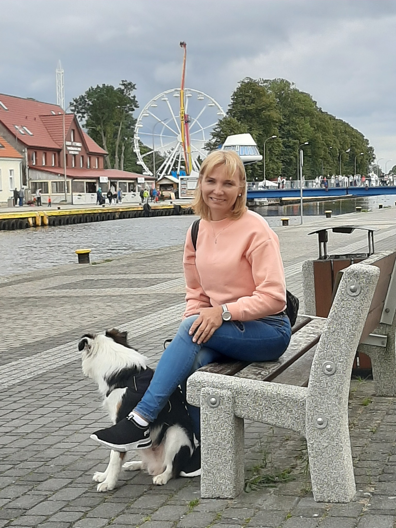 Uśmiechnięta kobieta w różowym swetrze i jeansach siedzi na ławce nad rzeką z psem rasy border collie u jej stóp, w tle widoczny diabelski młyn i budynki.
