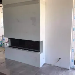 Nowoczesny, minimalistyczny kominek z długim, poziomym paleniskiem, obudowany płytami gipsowo-kartonowymi w trakcie wykończenia, widoczne łączenia płyt wymagające szpachlowania, szare płytki...