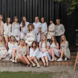 Grupowe zdjęcie kobiet, w tym jedna w ciąży, na baby shower w plenerze, z dekoracjami z suszonych traw i lustrem z napisem 'Baby Shower'.