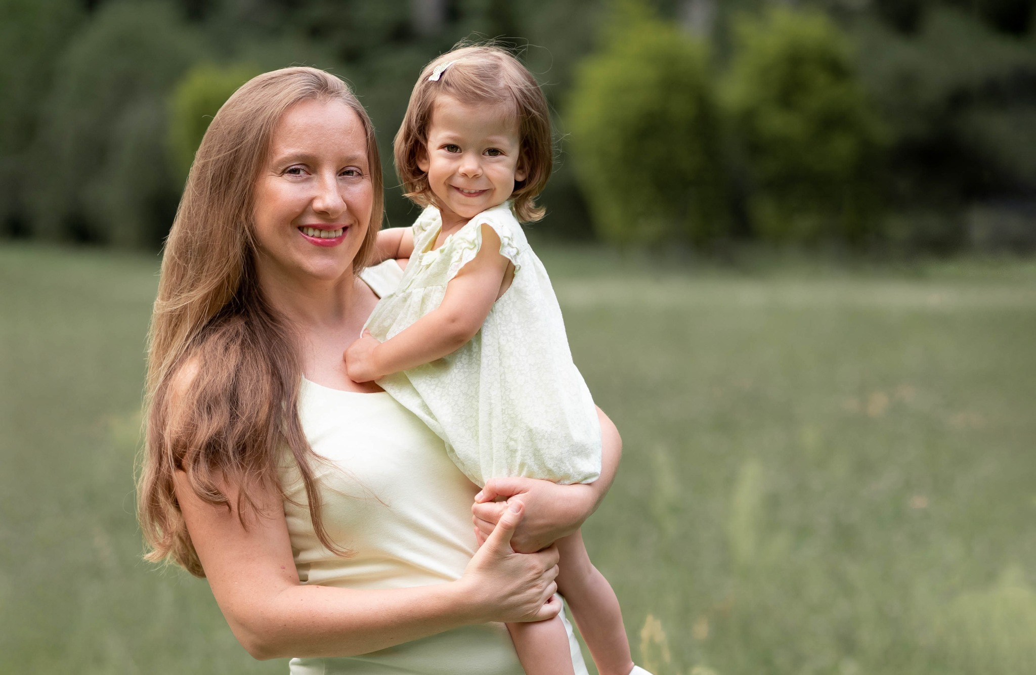 Uśmiechnięta mama trzymająca na rękach małą córeczkę w letniej sukience, na tle zieleni parku.