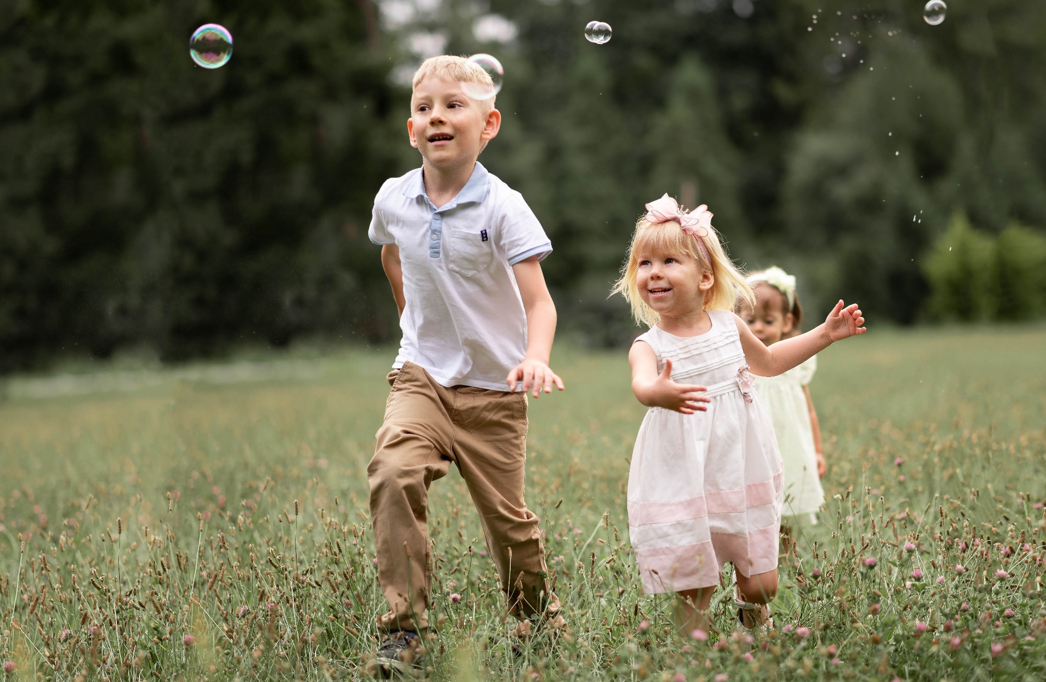 Radosny blond chłopiec i dwie dziewczynki w sukienkach biegają po łące w pogoni za bańkami mydlanymi, w tle zieleń drzew