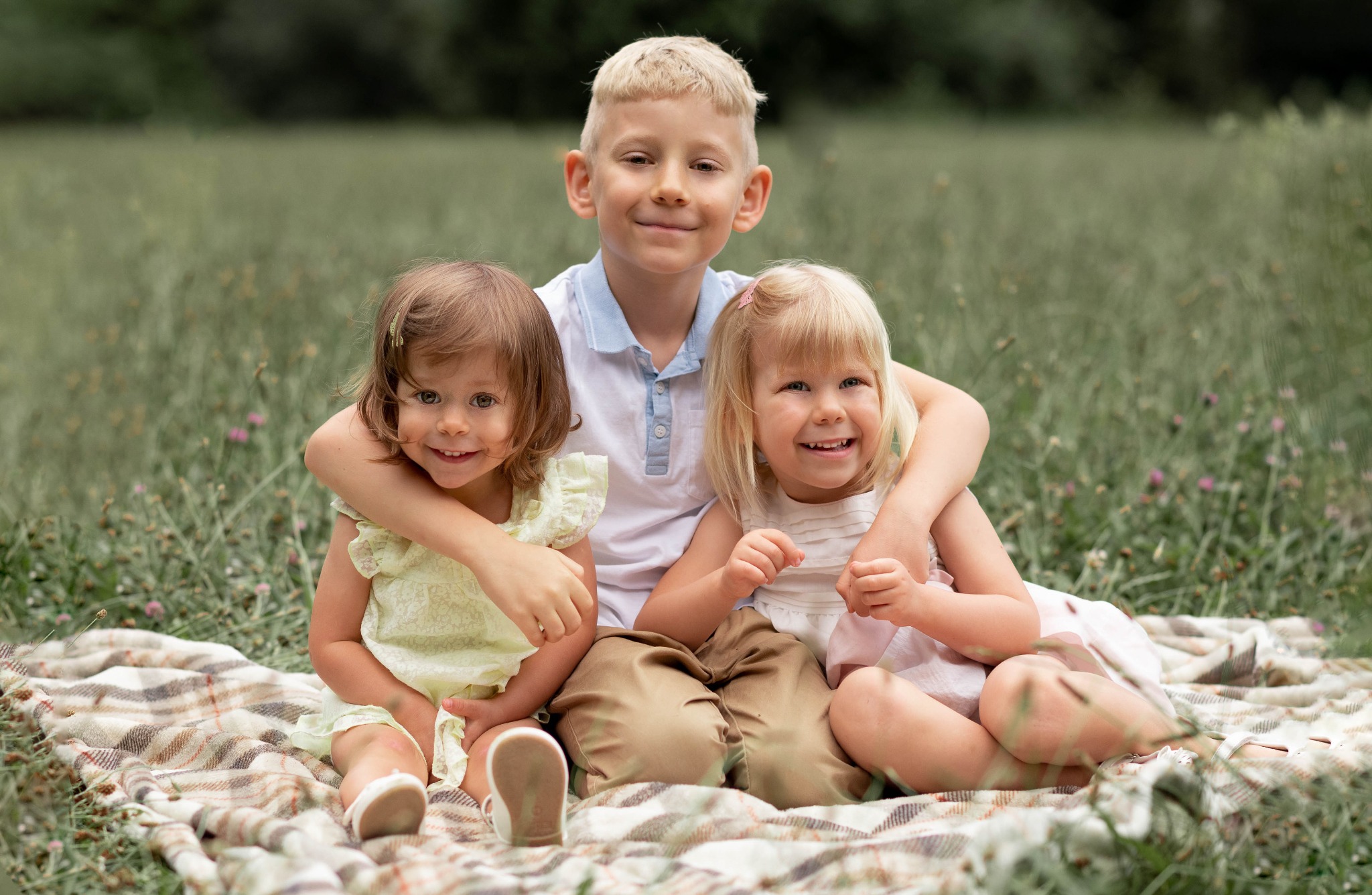Uśmiechnięty chłopiec obejmuje dwie małe dziewczynki siedzące na pledzie w plenerze, z rozmyciem w tle.