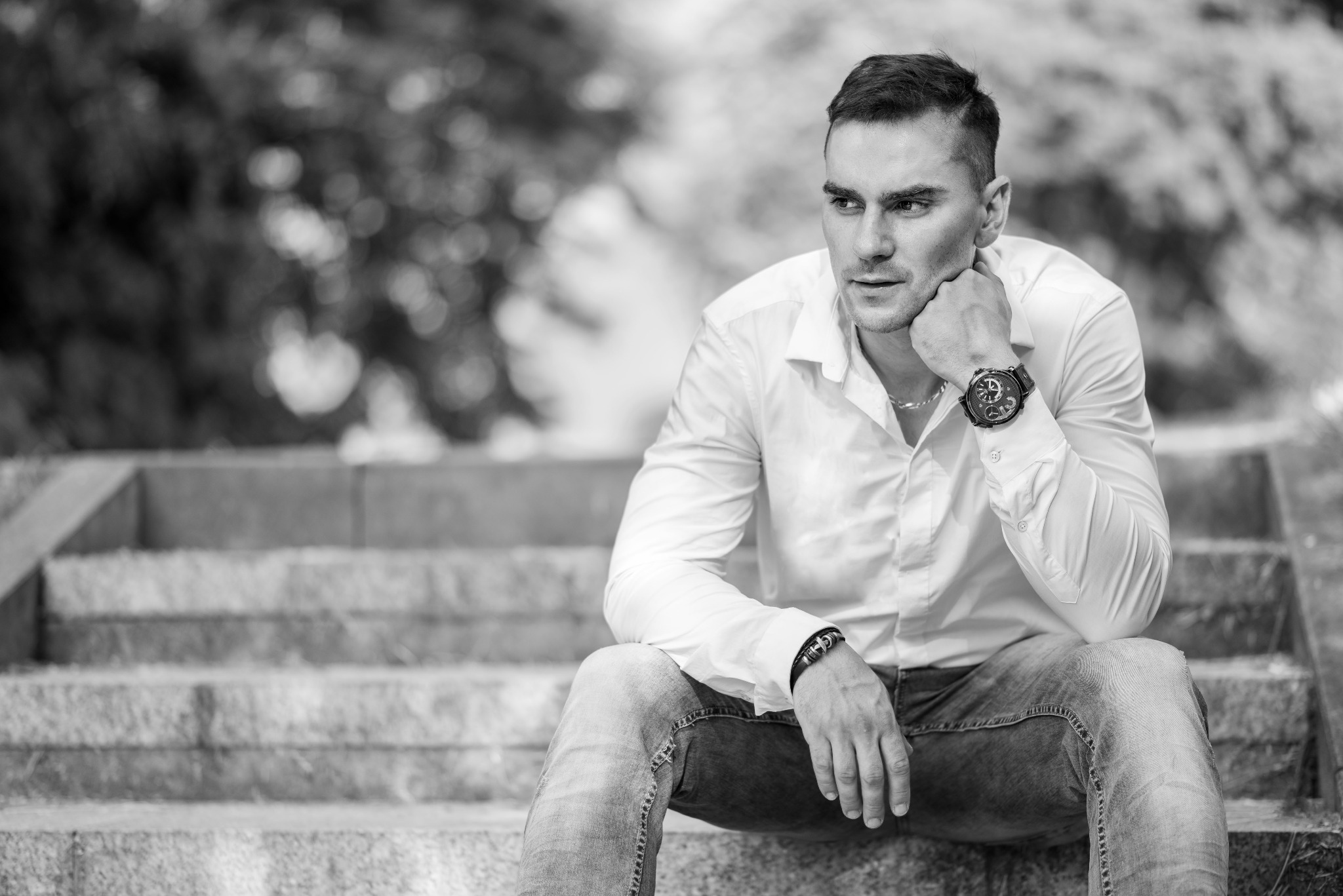 Czarno-białe zdjęcie portretowe mężczyzny w białej koszuli i jeansach, siedzącego na betonowych schodach z zamyśloną miną, z widocznym zegarkiem na ręce i bransoletką na nadgarstku, w tle rozmyta...