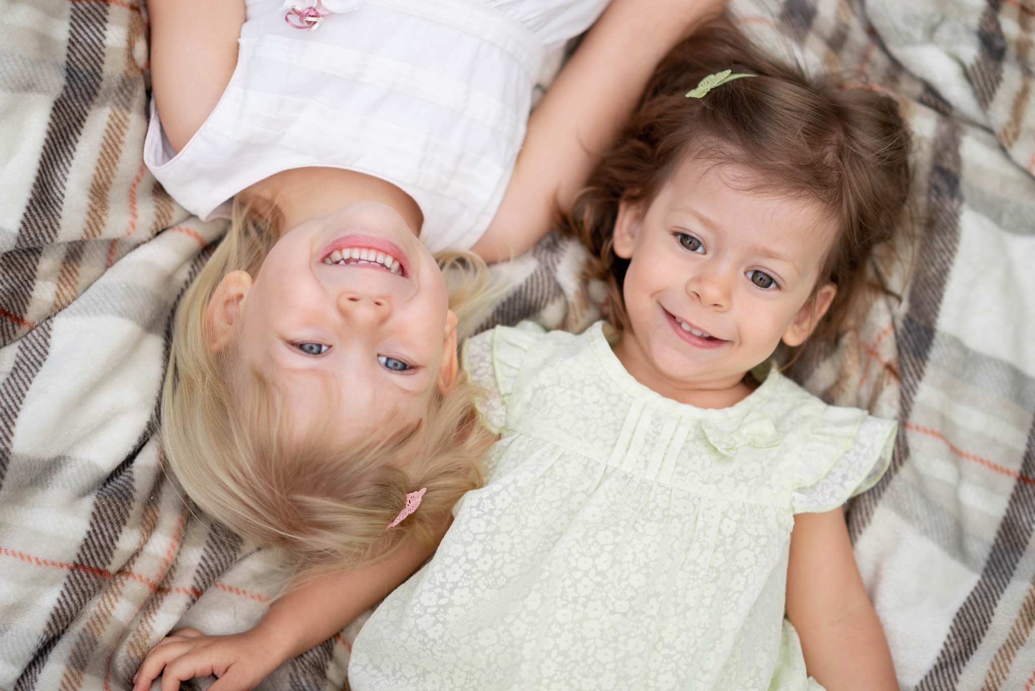 Dwie uśmiechnięte dziewczynki, blondynka i brunetka, leżą obok siebie na pledzie w kratę, ubrane w letnie sukienki z delikatnymi wzorami.
