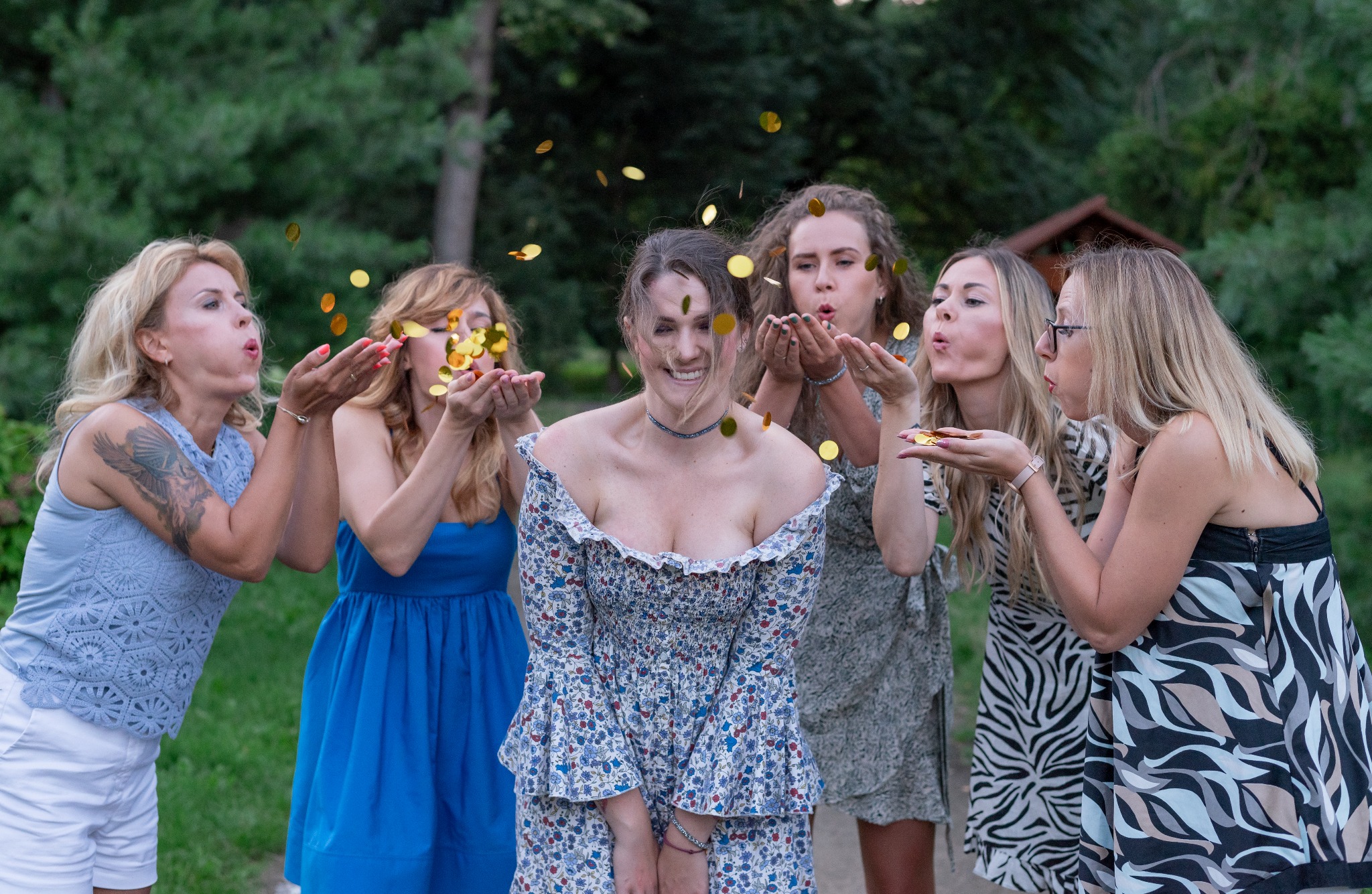 Kobieta w kwiecistej sukience uśmiecha się, otoczona czterema przyjaciółkami dmuchającymi w jej stronę konfetti w kształcie złotych kółek na tle zieleni.