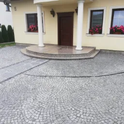 Podjazd do domu wyłożony szarą kostką granitową ułożoną w wachlarz, z wyznaczonym chodnikiem z większych płyt i obrzeżem z ciemnej kostki, prowadzący do schodów wejściowych z kolumnami.