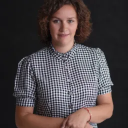 Portret uśmiechniętej kobiety z kręconymi włosami, ubrana w sukienkę w czarno-białą kratę, pozująca na ciemnym tle.