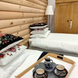 Sypialnia w stylu rustykalnym z drewnianymi ścianami, łóżko z białą pościelą i poduszkami z ludowymi wzorami, na tacy zestaw do herbaty w niebiesko-białe wzory.