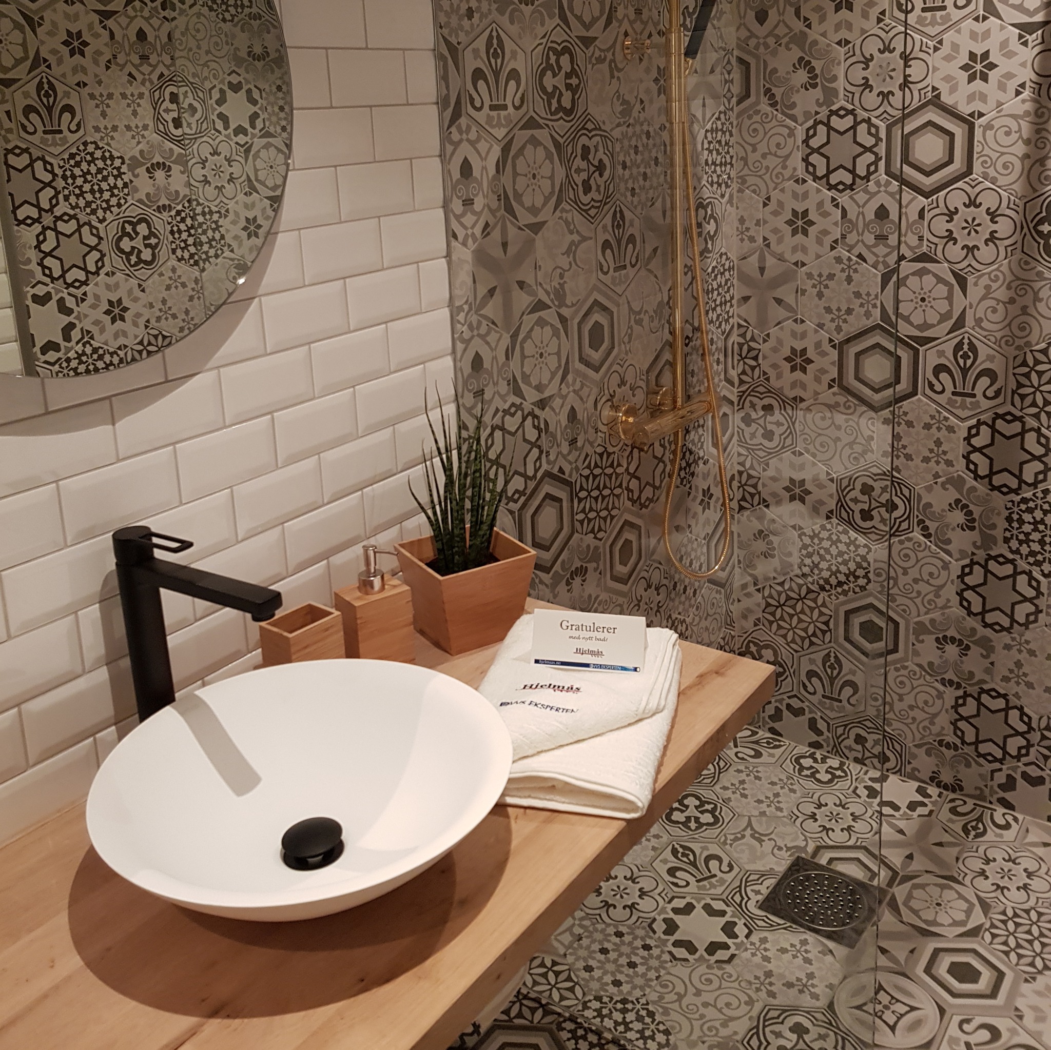 Nowoczesna łazienka z białą umywalką nablatową, czarną armaturą i drewnianym blatem, wykończona białymi kaflami typu metro oraz dekoracyjnymi szarymi płytkami heksagonalnymi w strefie prysznica.