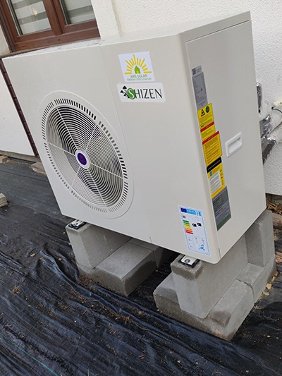 Biała pompa ciepła marki Shizen, umieszczona na betonowych bloczkach, z widocznymi naklejkami informacyjnymi i logo firmy Ars Solar, zainstalowana na zewnątrz budynku na czarnej folii.