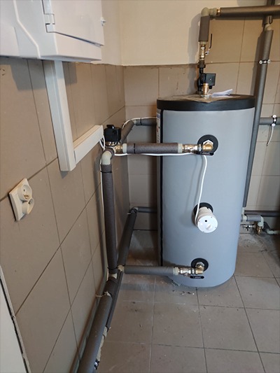 Szary zbiornik akumulacyjny ciepłej wody użytkowej z zaizolowanymi rurami, zamontowany w pomieszczeniu z szarą glazurą, widoczne przyłącza hydrauliczne i elektryczne.