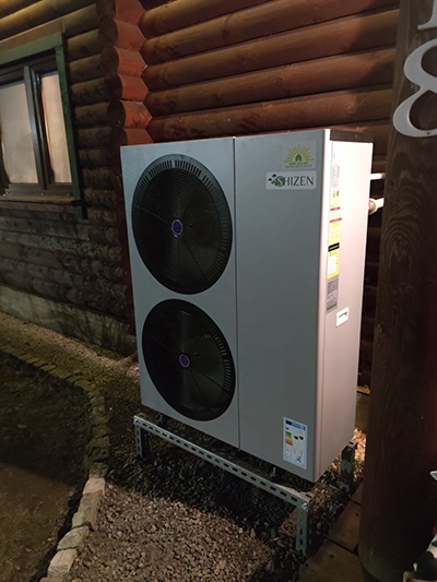 Zewnętrzna jednostka pompy ciepła marki Shizen, zamontowana na metalowej konstrukcji przy drewnianym domu z bali, widoczna naklejka z klasą energetyczną.