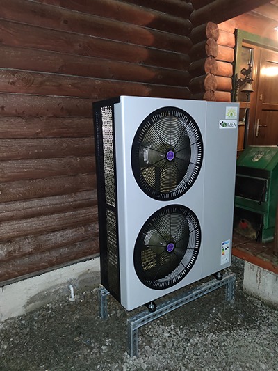 Szara pompa ciepła z dwoma wentylatorami, zamontowana na metalowej konstrukcji, stoi na żwirowym podłożu przed drewnianym domem w Olsztynie. Widoczne logo producenta.