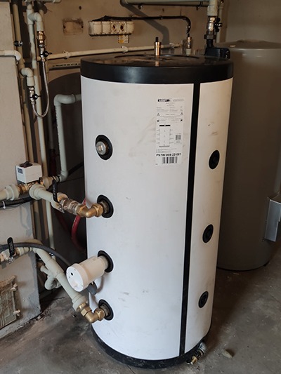 Biały, cylindryczny zbiornik akumulacyjny ciepła z podłączonymi rurami miedzianymi i plastikowymi w kotłowni. Widoczna etykieta z parametrami technicznymi na obudowie.