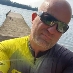 Mężczyzna w okularach przeciwsłonecznych i koszulce rowerowej marki Scott robiący selfie na tle drewnianego pomostu nad jeziorem w słoneczny dzień.