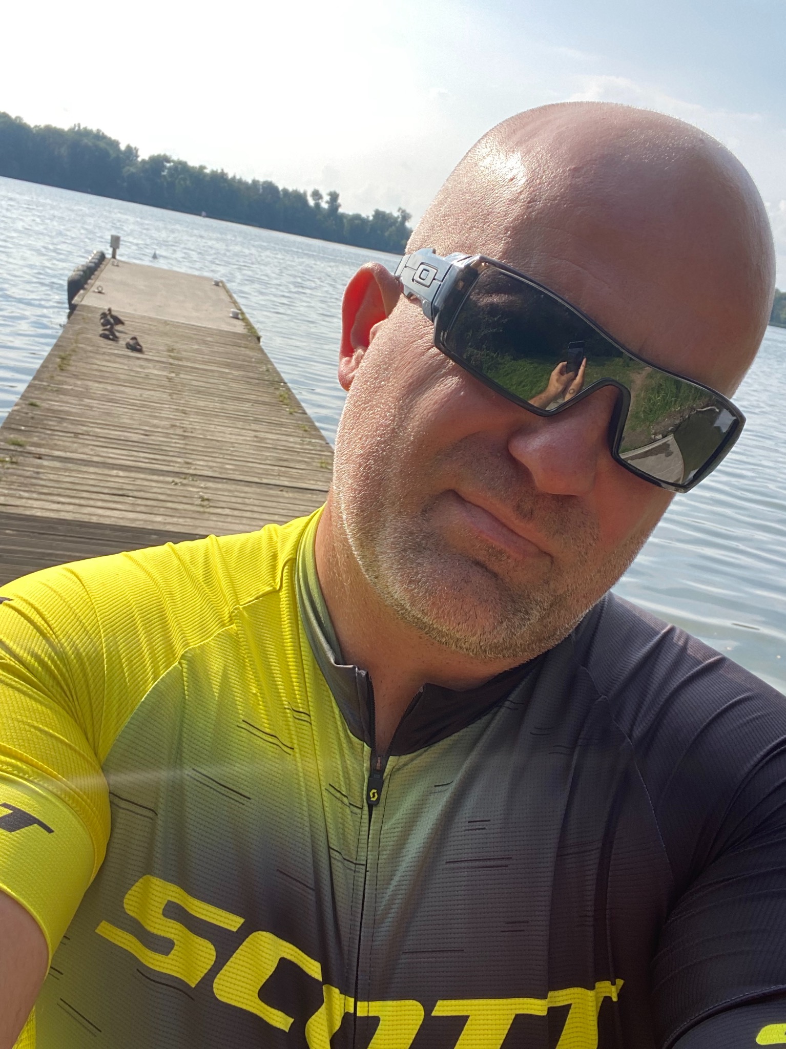 Mężczyzna w okularach przeciwsłonecznych i koszulce rowerowej marki Scott robiący selfie na tle drewnianego pomostu nad jeziorem w słoneczny dzień.