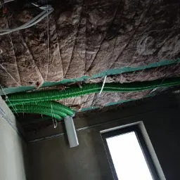 Montaż rekuperacji: zielone rury wentylacyjne pod sufitem z izolacją z wełny mineralnej, widoczne elementy mocujące i fragment okna.