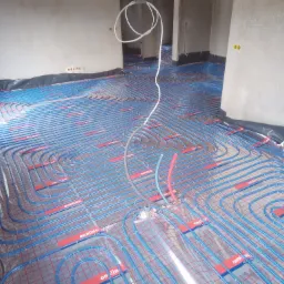 Rozłożone niebieskie rurki ogrzewania podłogowego na folii z czerwonymi paskami mocującymi, widoczne złącza i białe rurki doprowadzające, ściana z notatkami technicznymi w tle.