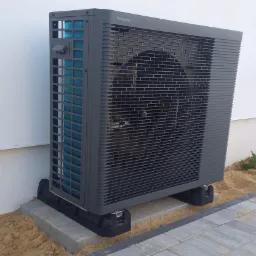 Szara, zewnętrzna jednostka pompy ciepła Panasonic, umieszczona na betonowych bloczkach i czarnych podkładkach antywibracyjnych, stoi przy białej ścianie budynku.