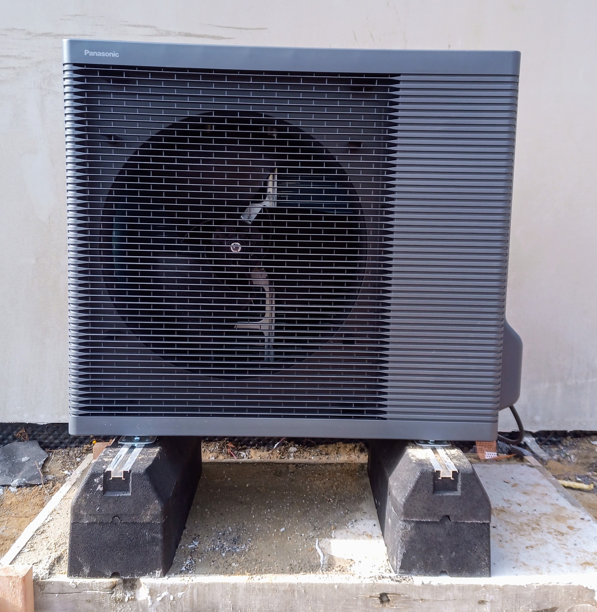 Zewnętrzna jednostka pompy ciepła Panasonic zamontowana na betonowych blokach z widocznymi elementami montażowymi, na tle jasnej ściany budynku.