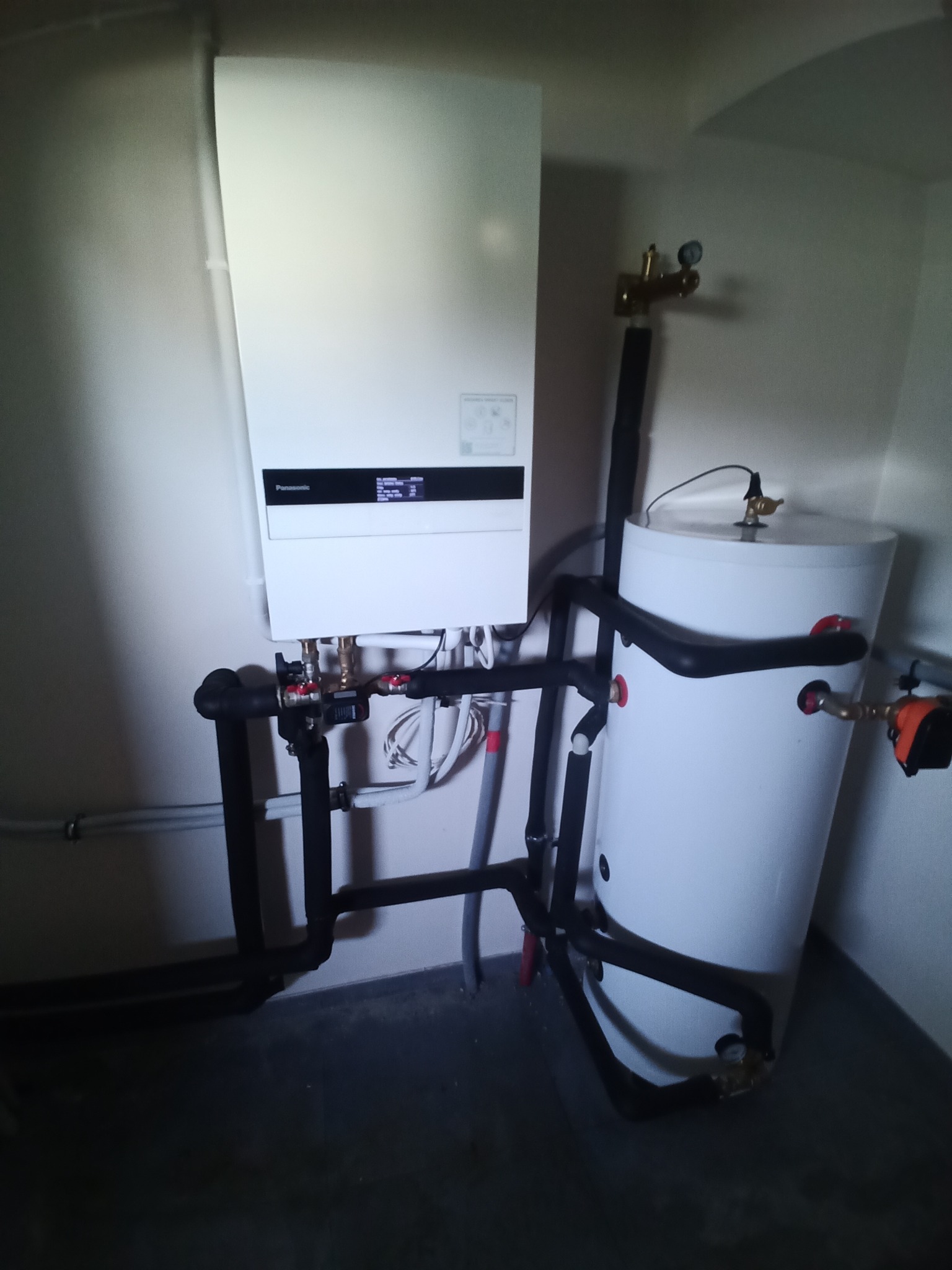 Biała pompa ciepła Panasonic Aquarea z zbiornikiem, połączona czarnymi rurami. Widoczne zawory i przewody. Ujęcie w rogu pomieszczenia.