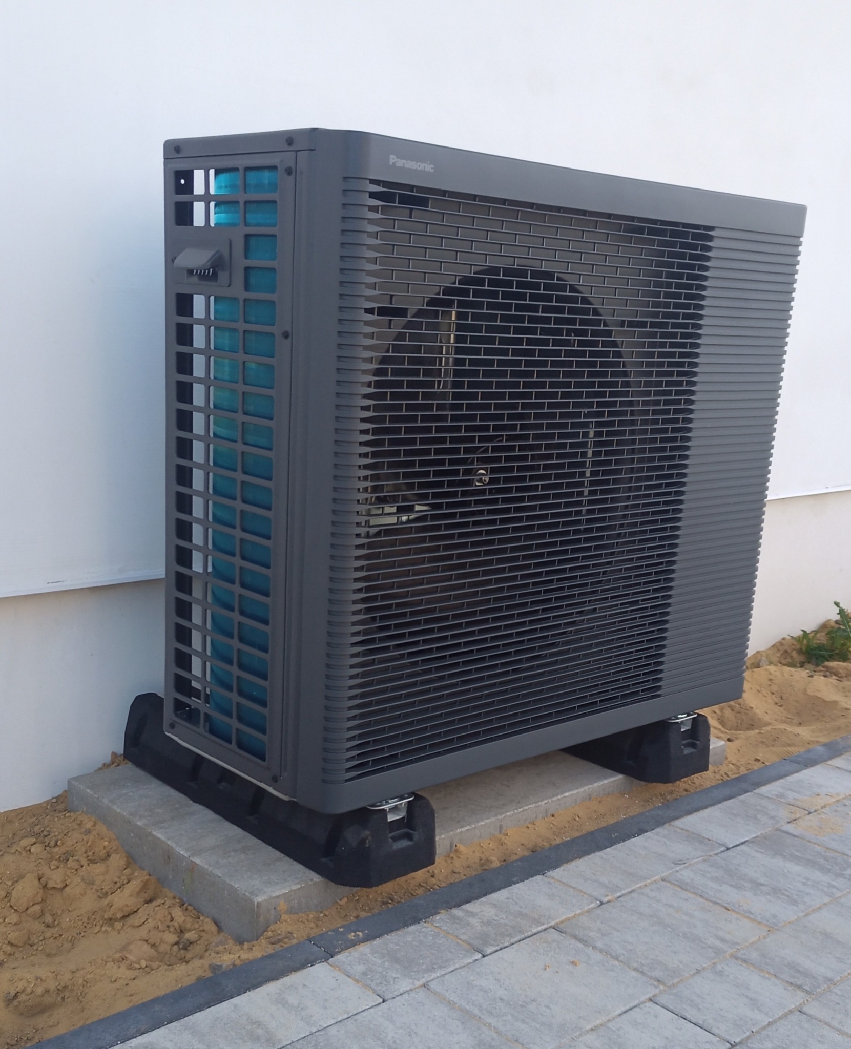 Szara, zewnętrzna jednostka pompy ciepła Panasonic, umieszczona na betonowych bloczkach i czarnych podkładkach antywibracyjnych, stoi przy białej ścianie budynku.