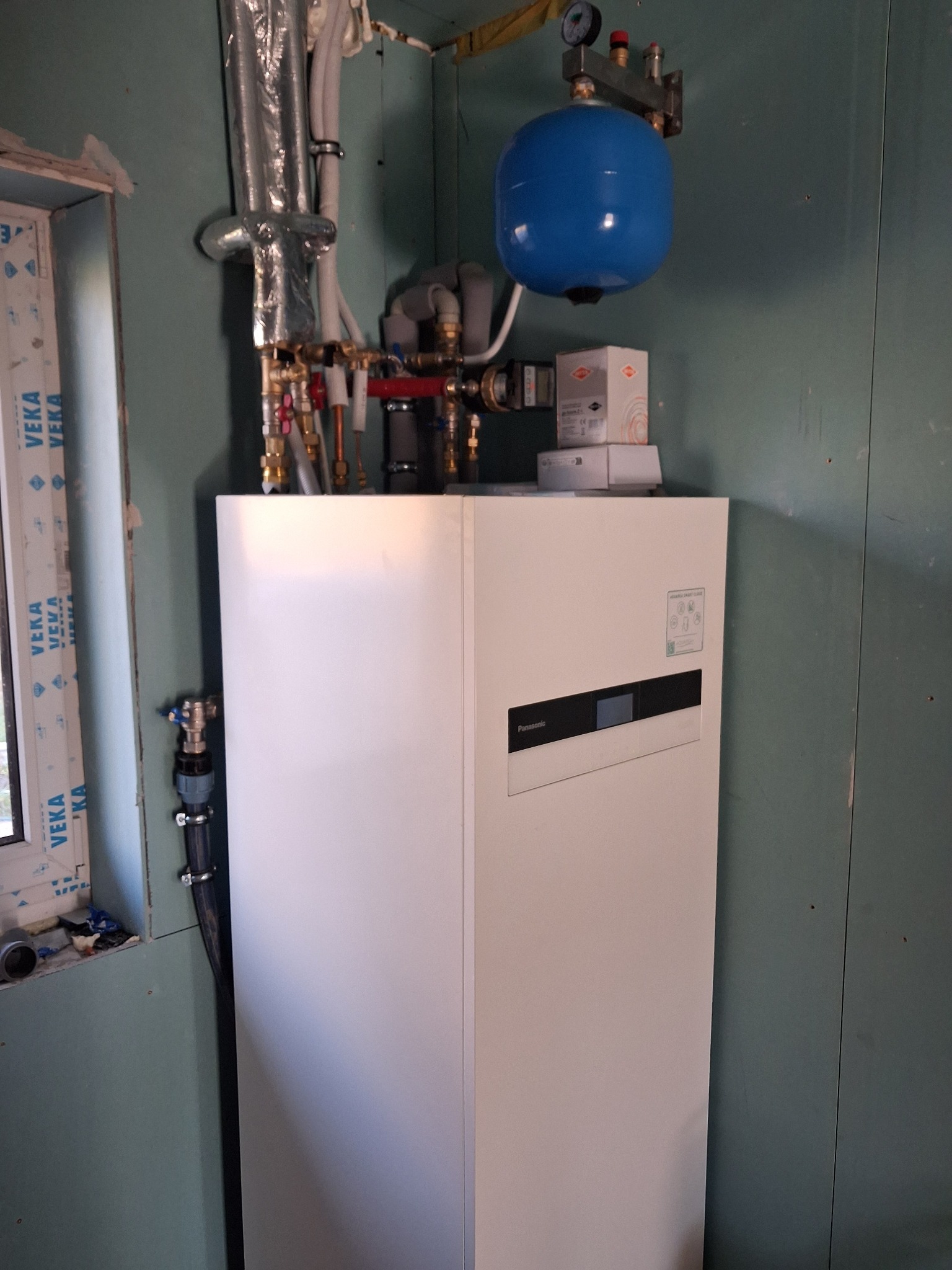 Biała pompa ciepła Panasonic z niebieskim zbiornikiem wyrównawczym i instalacją hydrauliczną w pomieszczeniu z oknem. Widoczne rury i zawory.