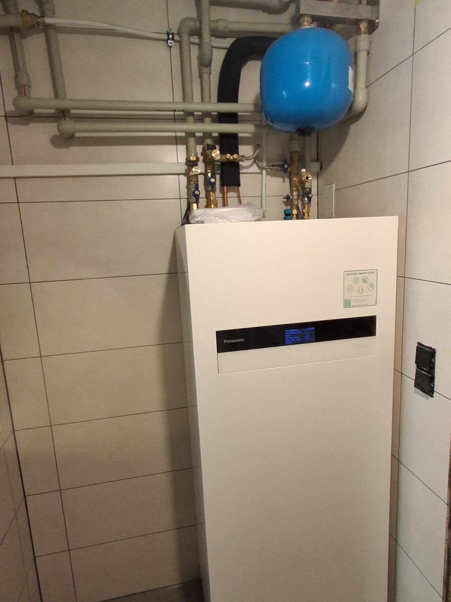 Biała pompa ciepła Panasonic Aquarea Smart Cloud z niebieskim zbiornikiem wyrównawczym i instalacją hydrauliczną w pomieszczeniu wyłożonym jasnymi płytkami.