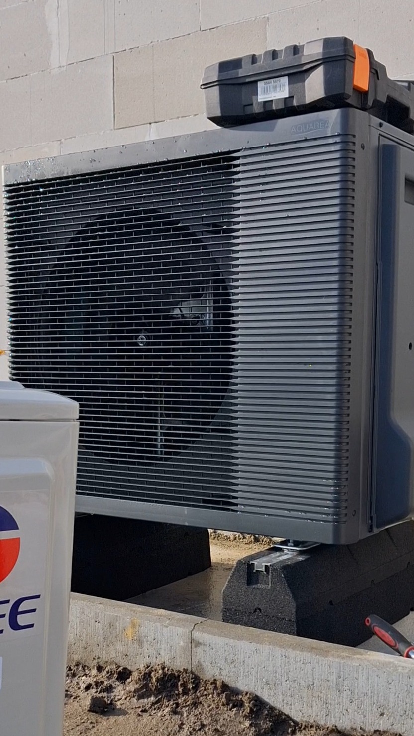 Szara pompa ciepła Aquarea na czarnych podkładkach, z skrzynką narzędziową na górze, stoi na zewnątrz budynku. Widoczny fragment białego zbiornika.