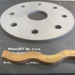 Obrabiany metalowy element w kształcie falistej listwy z mosiądzu oraz okrągły element z otworami na czarnym podłożu z nadrukiem WaterJET.